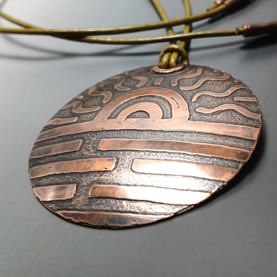 Up close photo of large etched copper pendant with sun motif on long adjustable leather cord. Displayed on grey background.