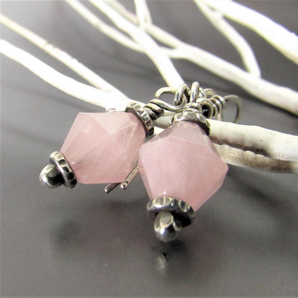 Close up of pink quarts earrings showing detail of the metalwork on the bottom, displayed against a white branch on a gray background. 