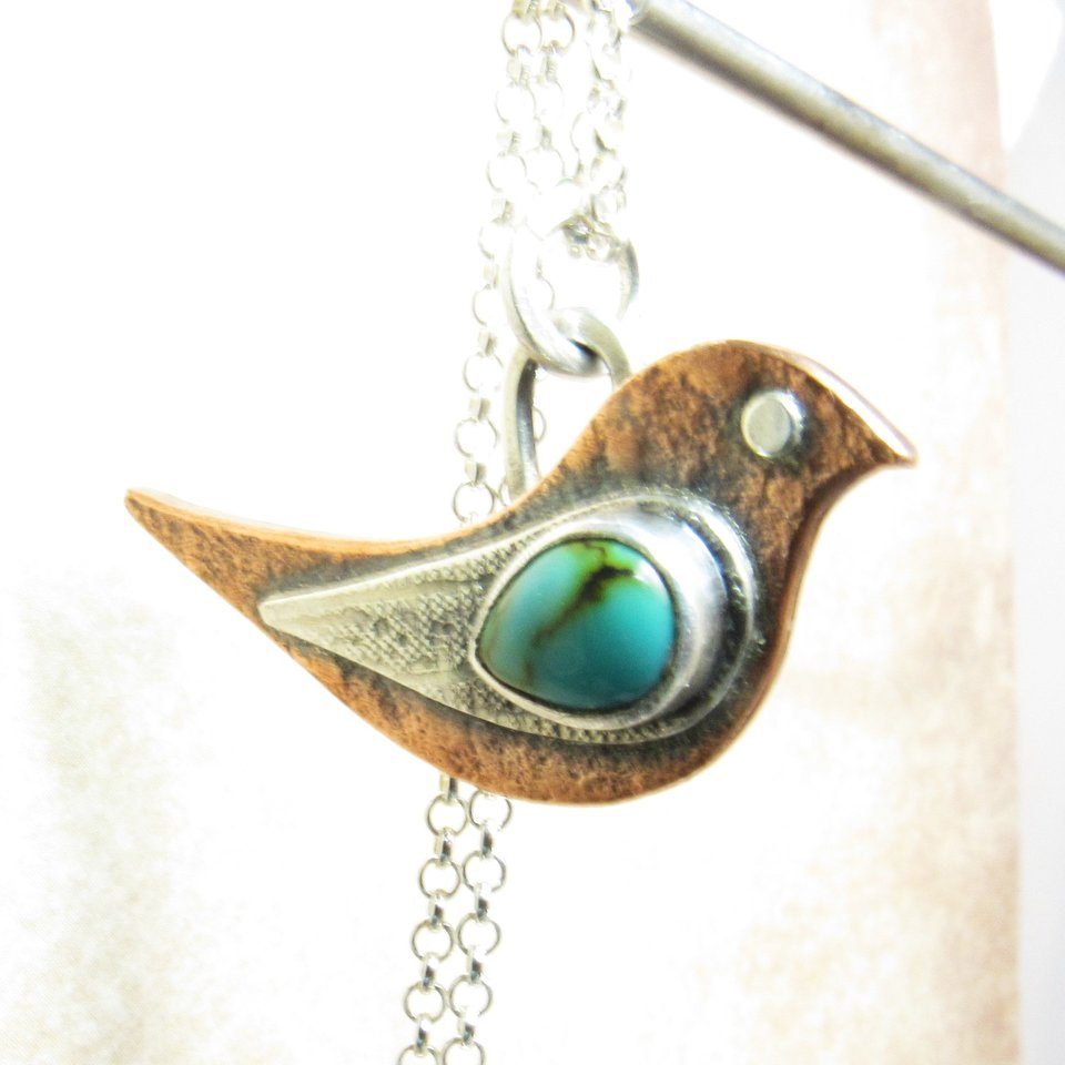 Bird pendant necklace in mixed metals with turquoise. Suspended from stand.