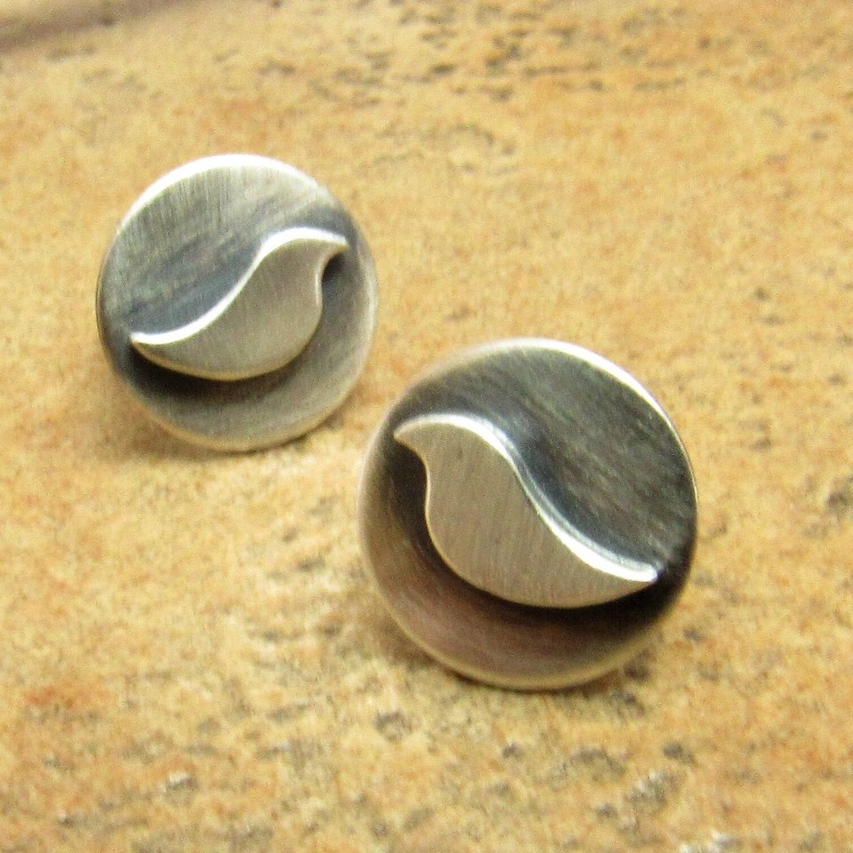 Front view of a pair of silver earrings with a bird design. The earrings are round and have a brushed finish displayed on sand color tile background.