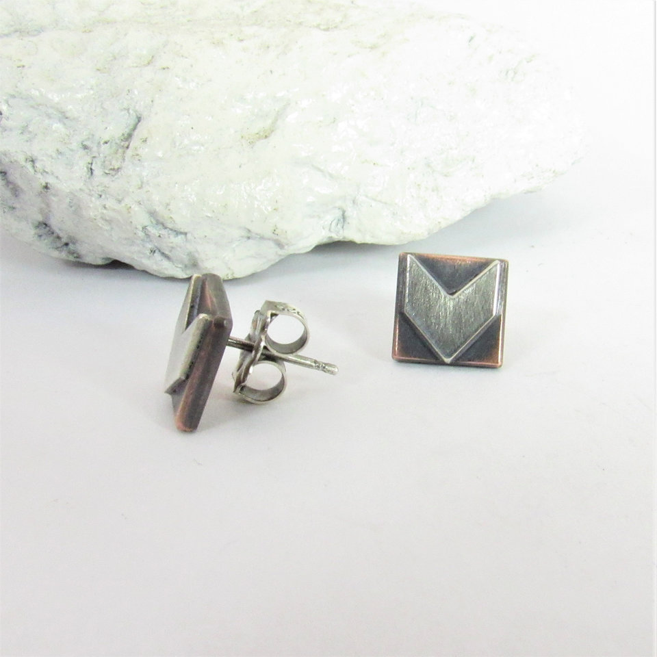 May include: A pair of silver and copper stud earrings with a geometric arrow design. The earrings are square shaped with a brushed silver arrow in the center. Clutch backs included. Displayed against rock on white background.
