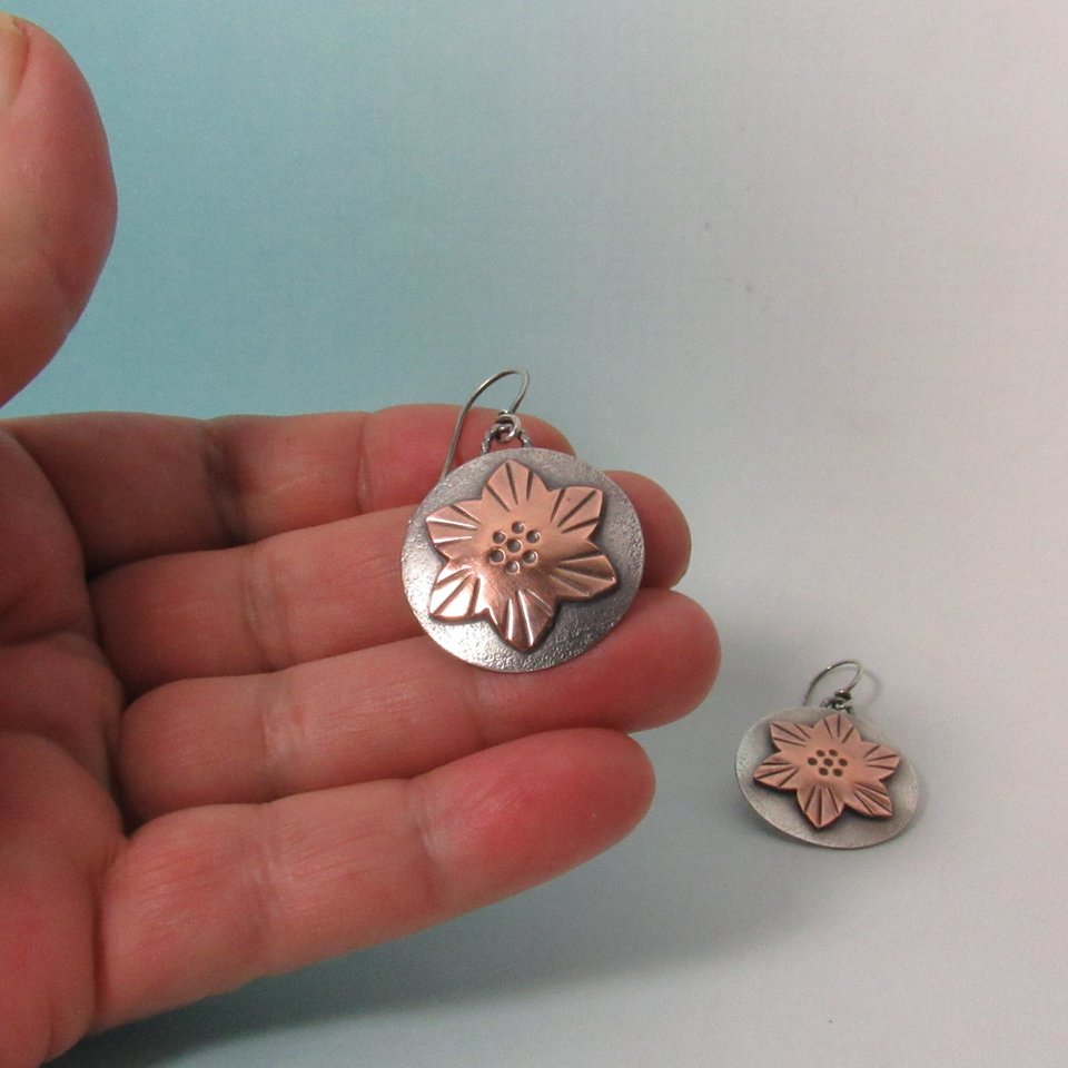 Copper and sterling silver earrings with a flower design. The copper flower is layered atop the sterling disk. Lightweight and measure 1.5" tall and 1" wide. Artisan jewelry to treasure. Displayed in hand against blue background.