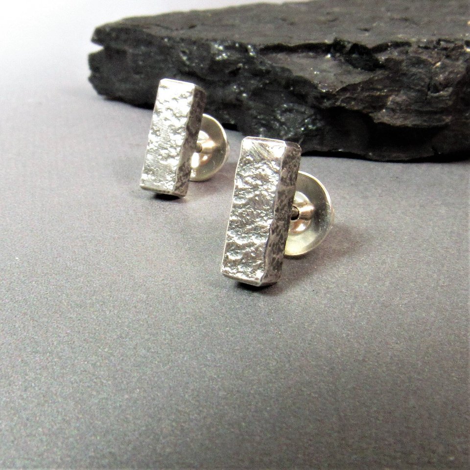 A pair of silver bar stud earrings with a textured finish. The earrings are on a gray surface with dark rock.