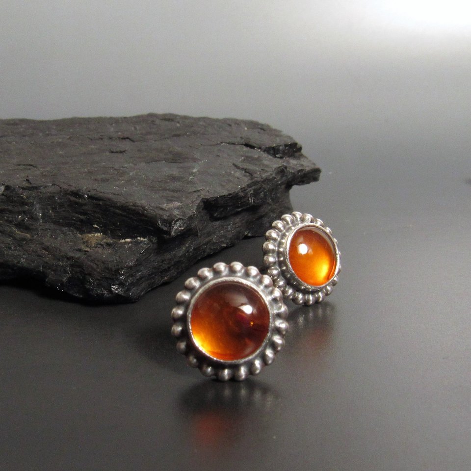 A pair of silver post back earrings with amber gemstone cabochons set in a beaded bezel. Displayed on dark rock and background