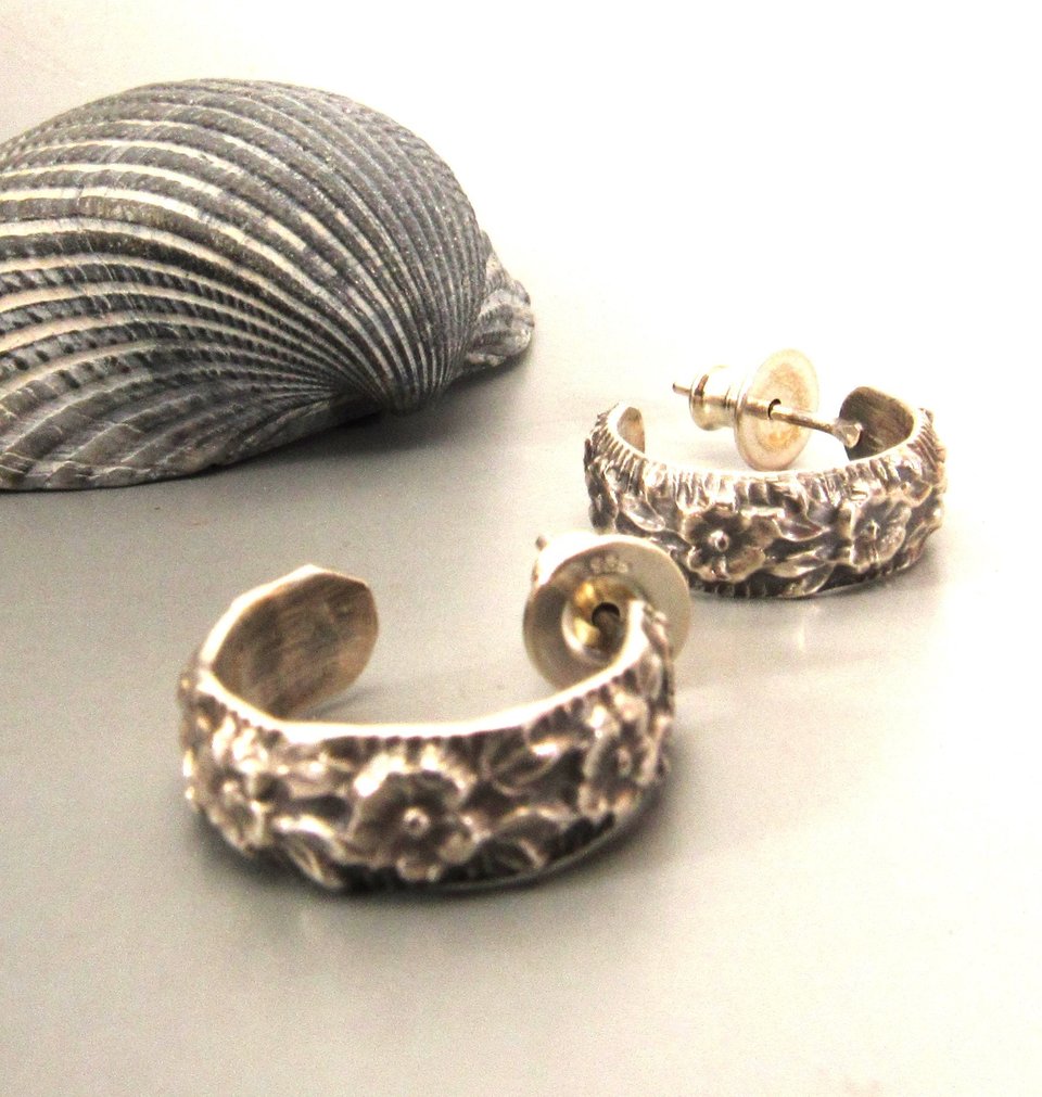 A pair of small silver stud hoop earrings with a floral design. The earrings are made of sterling silver and have a polished finish.