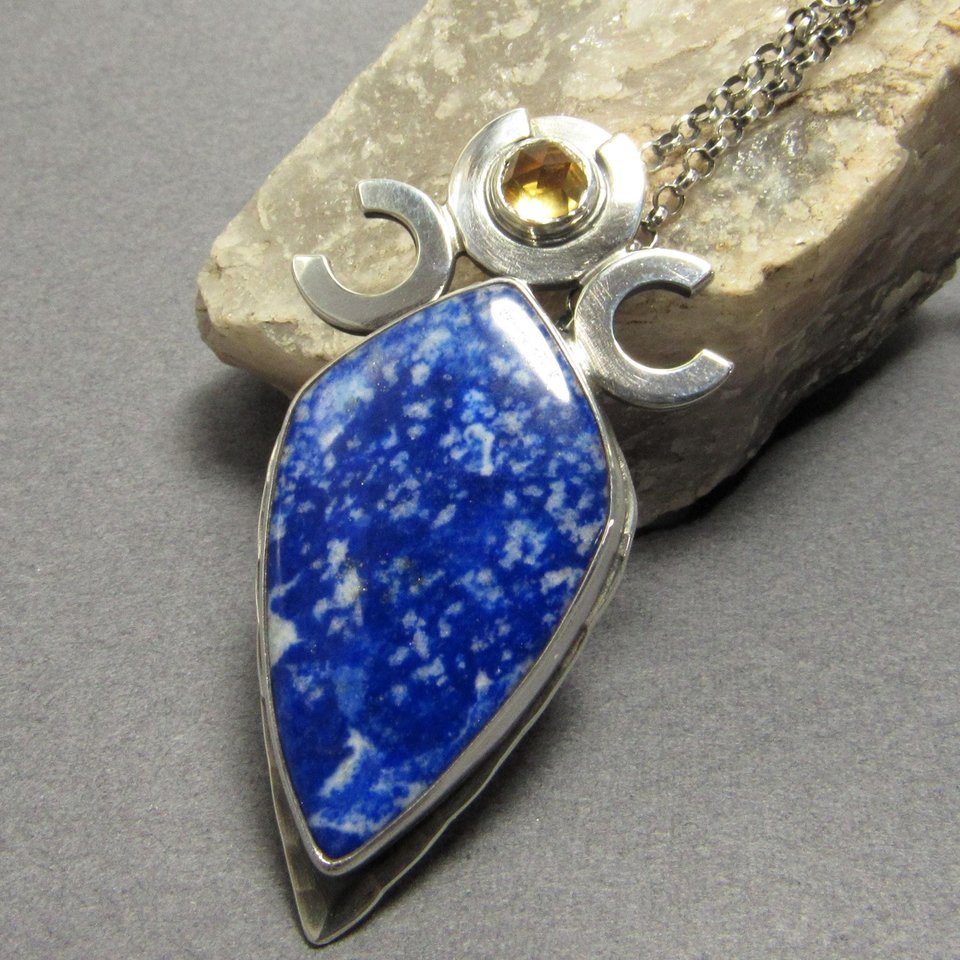 Triple goddess / moon pendant necklace with a faceted citrine and large lapis lazuli cabochon crafted from argentium sterling silver, fine silver. Pendant measures 2 inches from the top and is 7/8 inch across widest part and is suspended on a 18 inch sterling silver chain. Displayed against rock on grey background.