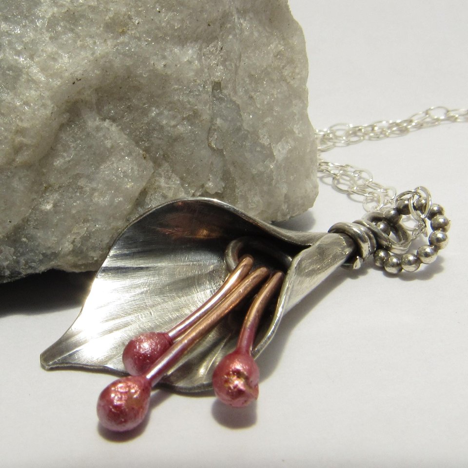A silver and copper pendant necklace with a lily flower design. The flower is made of silver and has three copper stems with pink tips. The necklace is attached to a silver chain with silver clasp.