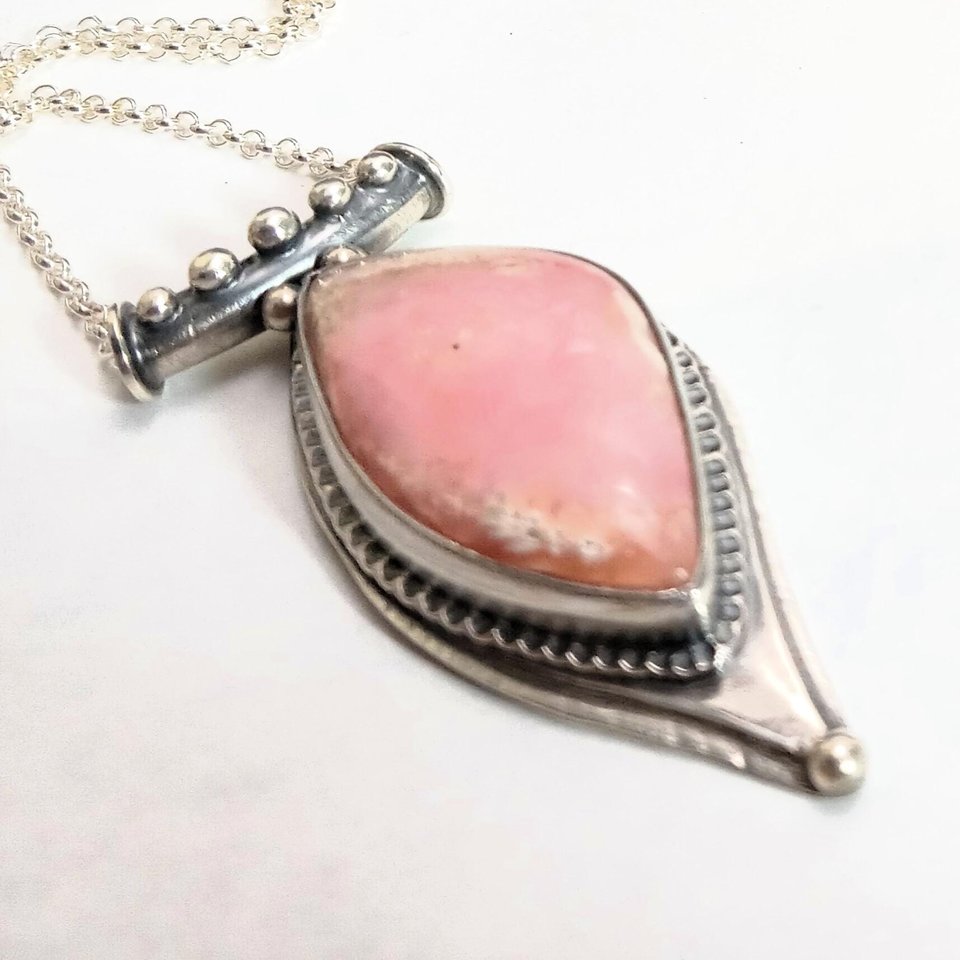 Soft elegance meets artisan craftsmanship in this one of a kind handcrafted pendant featuring a beautifully pastel pink opal set in textured Argentium® sterling silver. The unique, petal-shaped stone in a petal shaped setting radiates gentle warmth in a soothing blush hue—perfect for those who adore subtle statement pieces with soulful charm. 2.25" long x 7/8" wide.