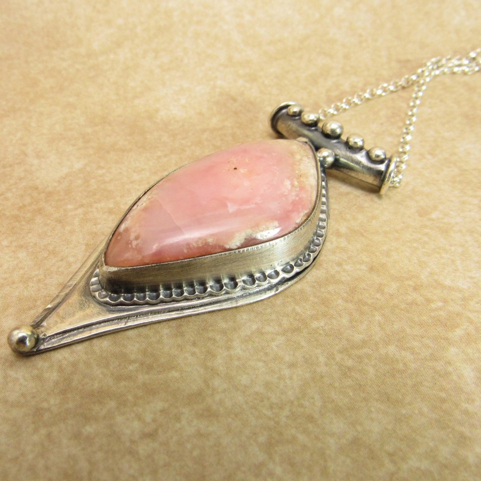 Soft elegance meets artisan craftsmanship in this one of a kind handcrafted pendant featuring a beautifully pastel pink opal set in textured Argentium® sterling silver. The unique, petal-shaped stone in a petal shaped setting radiates gentle warmth in a soothing blush hue—perfect for those who adore subtle statement pieces with soulful charm. 2.25" long x 7/8" wide.