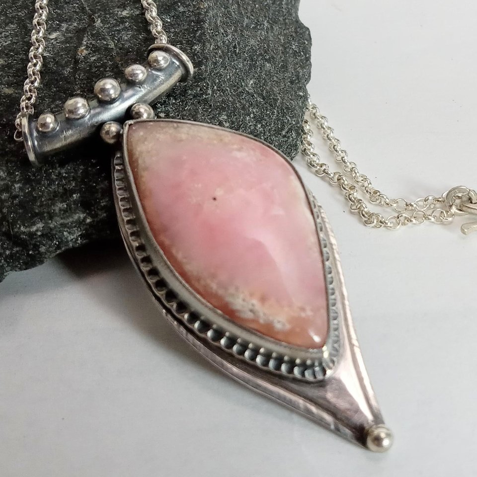 Soft elegance meets artisan craftsmanship in this one of a kind handcrafted pendant featuring a beautifully pastel pink opal set in textured Argentium® sterling silver. The unique, petal-shaped stone in a petal shaped setting radiates gentle warmth in a soothing blush hue—perfect for those who adore subtle statement pieces with soulful charm. 2.25" long x 7/8" wide.