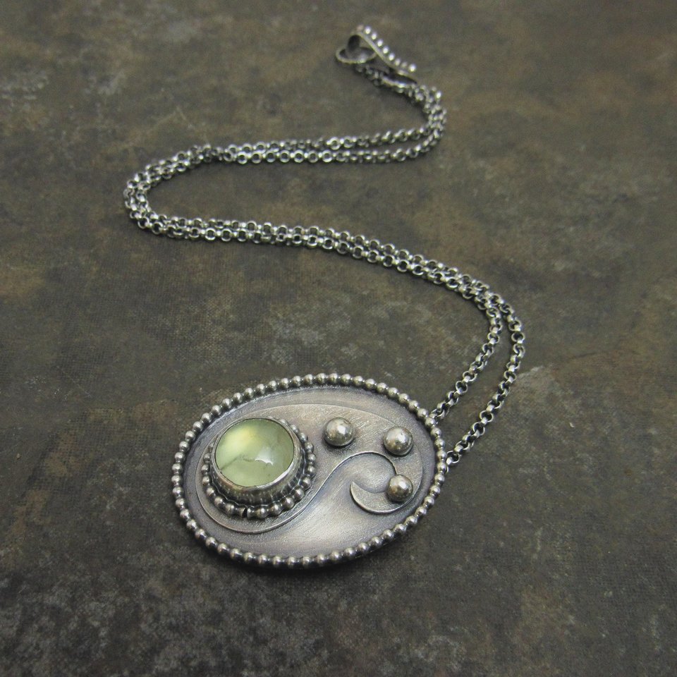 Green prehnite cabochon and argentium sterling silver pendant necklace on dark stone colored background with chain and clasp trailing in the background.