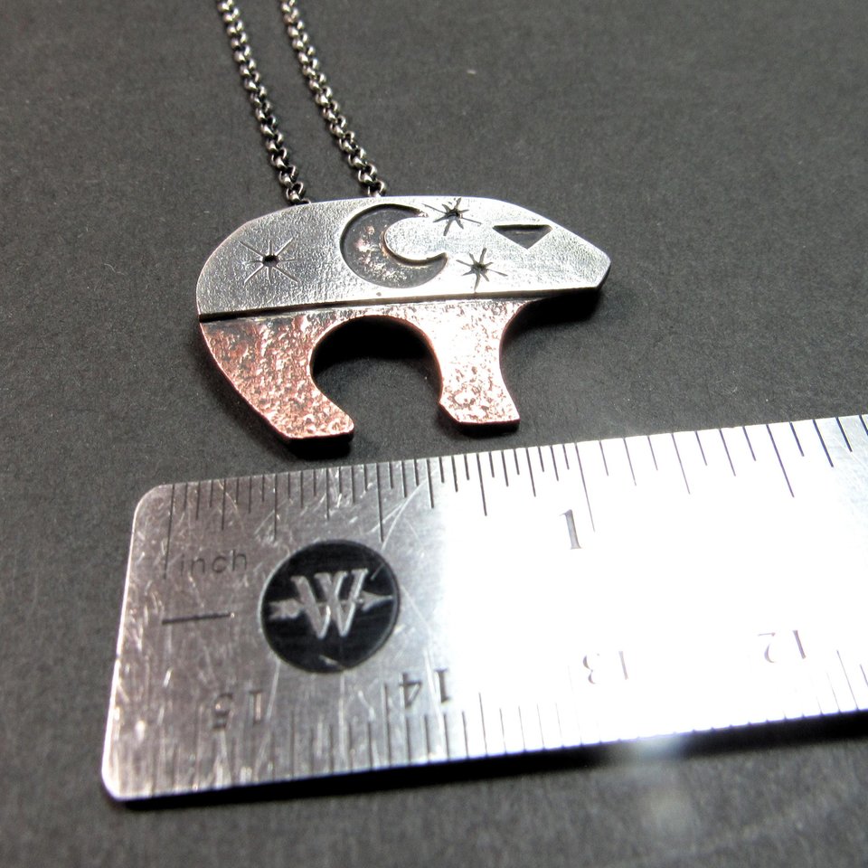 Rustic handmade mixed metal bear necklace with a sterling silver sky design, crescent moon, eye, star cutouts, and textured copper base, finished with oxidation and polishing for depth and contrast. Displayed on a grey background against ruler to show width. 18" SS chain and handmade findings included.