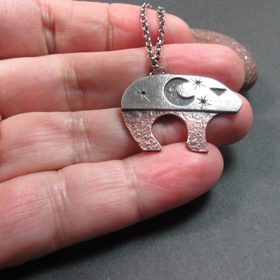 Artisan bear pendant in copper and sterling silver, showcasing a layered design with celestial symbols, smooth silver bail, and an 18 inch sterling silver chain, bold yet earthy in style. Displayed in hand with rock and grey background.