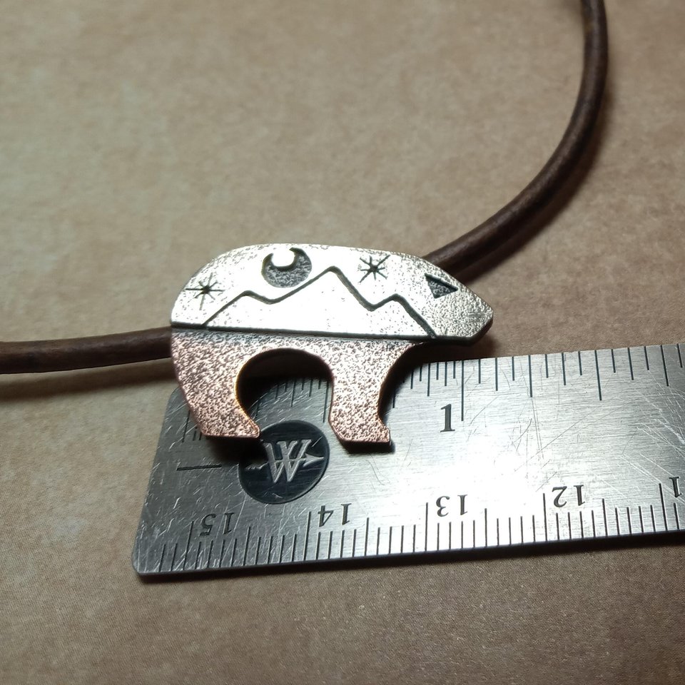 Bear pendant by Mocahete against ruler to show size. Bear pendant with the top half sculpted in silver with moon and star cutouts and the lower body in richly textured copper. The pendant is suspended on a sturdy 18-22 inch leather cord finished with our handmade Argentium silver findings for durability and comfort.Bear measures 1.25" long and is .75" tall. Displayed on ruler to show size.