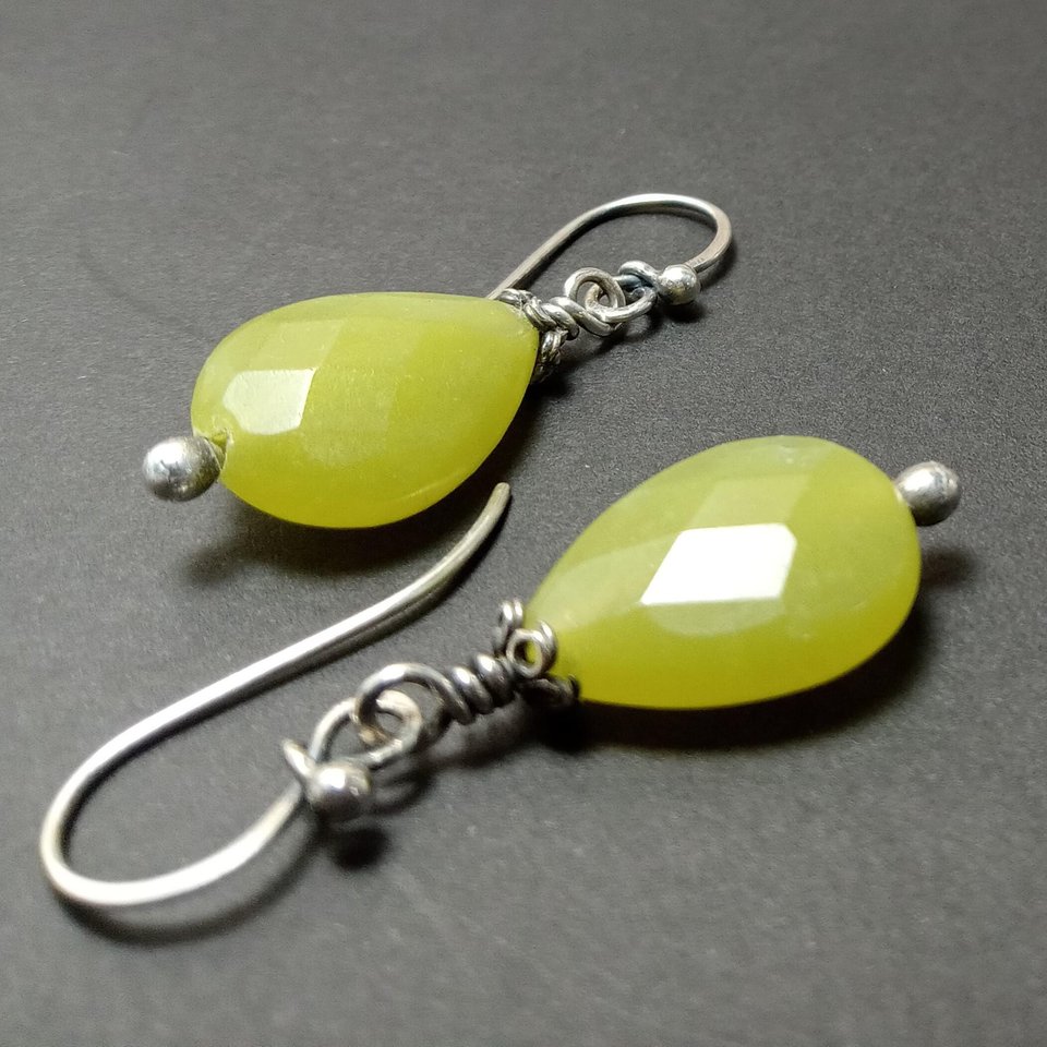 Close-up of handcrafted green faceted serpentine teardrop earrings with sterling silver bead and cap accents, hanging from artisan-made Argentium ear wires on a dark background.
