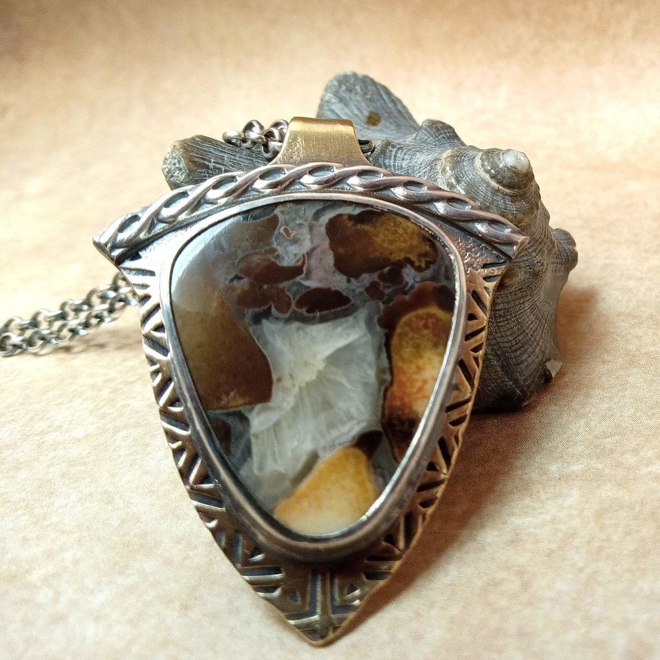 A handcrafted mixed-metal pendant necklace featuring a triangular Cobra Jasper stone with earthy coffee, golden yellow, and cream tones set in a fine silver bezel on a bronze base, with sterling silver patterned wire accents, shown on a sturdy silver chain against a natural shell background.