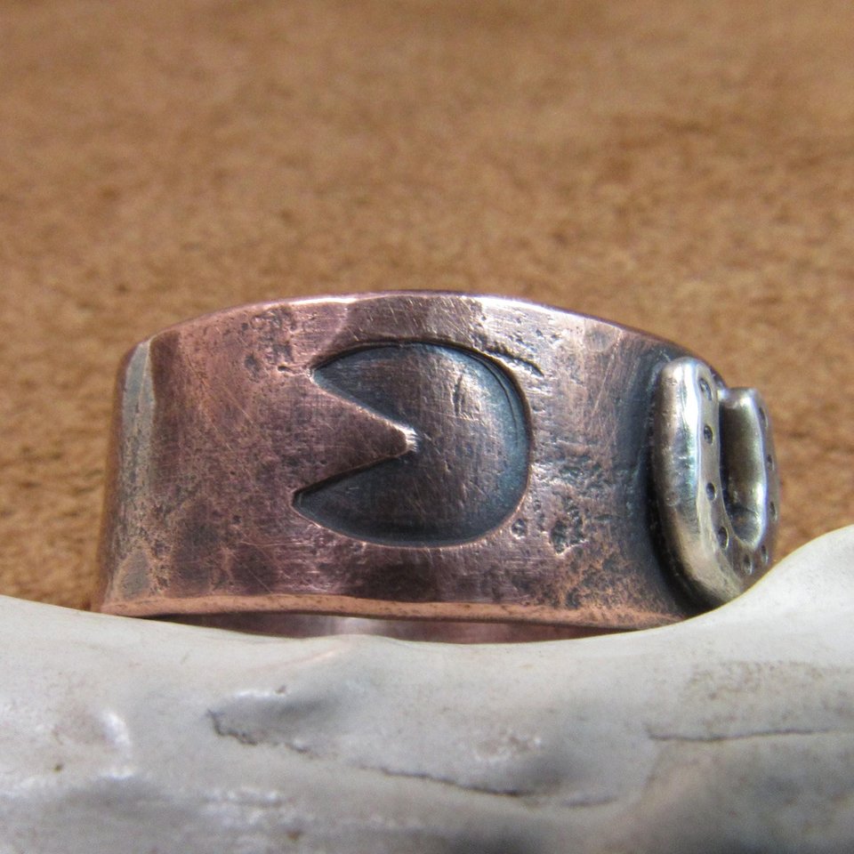 Wide rustically textured copper band with sterling silver horseshoe and horse hoof print stamp motif. Ring has been oxidized and polished giving it a deep and rich patina, and is photographed on grey background propped on a deer antler with the hoof stamping facing forward. Band is made in whole and half sizes from 6-16.