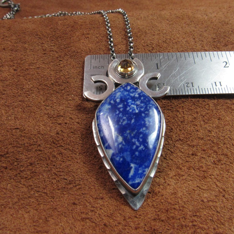 Vertical view of Triple goddess / moon pendant necklace with a faceted citrine and large lapis lazuli cabochon crafted from argentium sterling silver, fine silver. Pendant measures 2 inches from the top and is 7/8 inch across widest part and is suspended on a 18 inch sterling silver chain. Displayed against ruler on brown leather.
