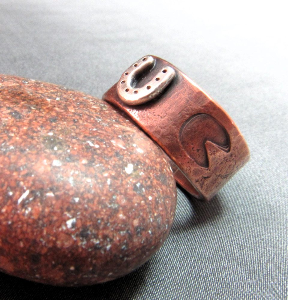 Wide rustically textured copper band with silver horseshoe and horse hoof print stamp motif. Ring has been oxidized and polished giving it a deep and rich patina, and is photographed on grey background propped on a red rock. Band is made in whole and half sizes from 6-16.