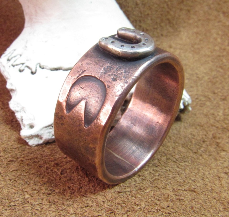 Wide rustically textured copper band with silver horseshoe and horse hoof print stamp motif. Ring has been oxidized and polished giving it a deep and rich patina, and is photographed on grey background stood up in front of a deer antler. Band is made in whole and half sizes from 6-16.