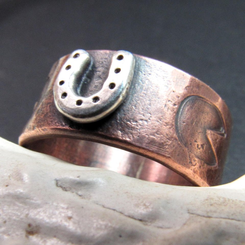 Wide rustically textured copper band with silver horseshoe and horse hoof print stamp motif. Ring has been oxidized and polished giving it a deep and rich patina, and is photographed on grey background propped on a deer antler. Band is made in whole and half sizes from 6-16.