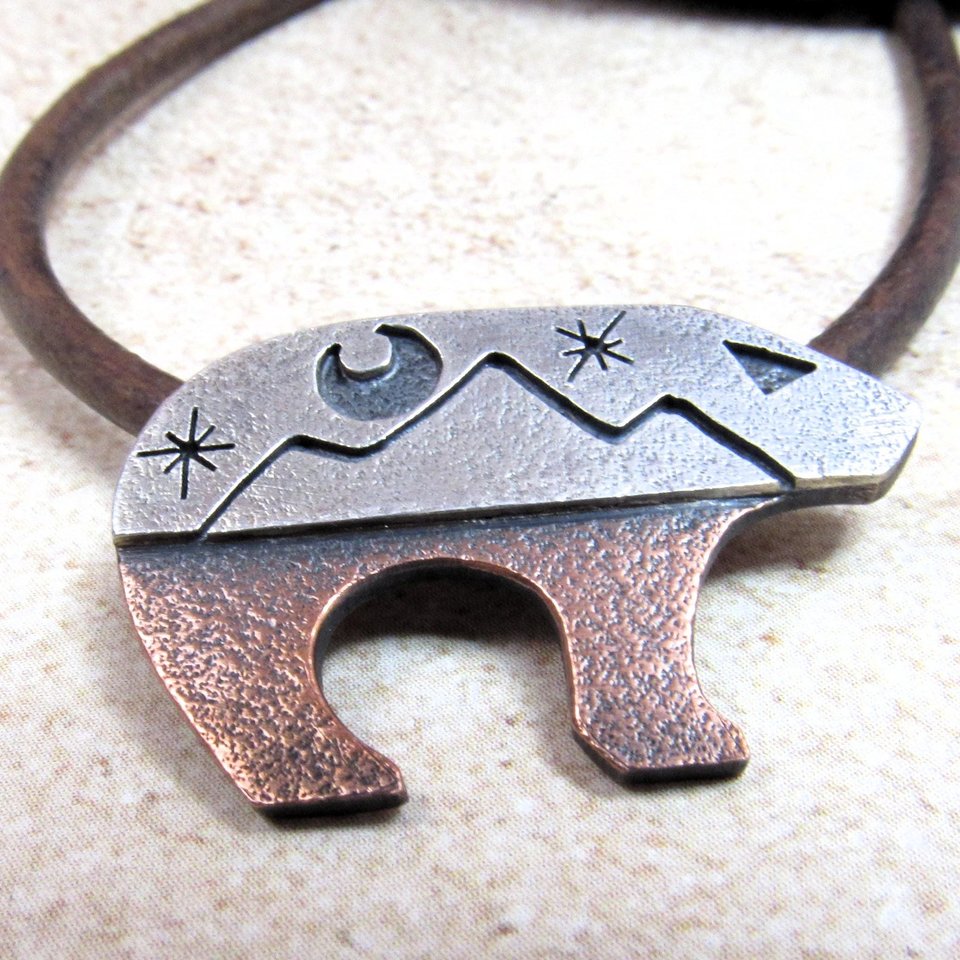Bear pendant with the top half sculpted in silver with moon and star cutouts and the lower body in richly textured copper, joined harmoniously through traditional metalsmith techniques. The pendant is suspended on a sturdy 18-22 inch leather cord finished with our handmade Argentium silver findings for durability and comfort.Bear measures 1.25" long and is .75" tall. Displayed on neutral background.