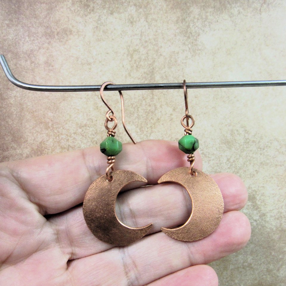 Handmade copper crescent moon dangle earrings with faceted green chrysoprase beads. Earrings measure 2 and 1/8" long and the moon is 3/8" wide at widest point.The ear wires are solid, pure copper. Displayed against hand hanging from stand.