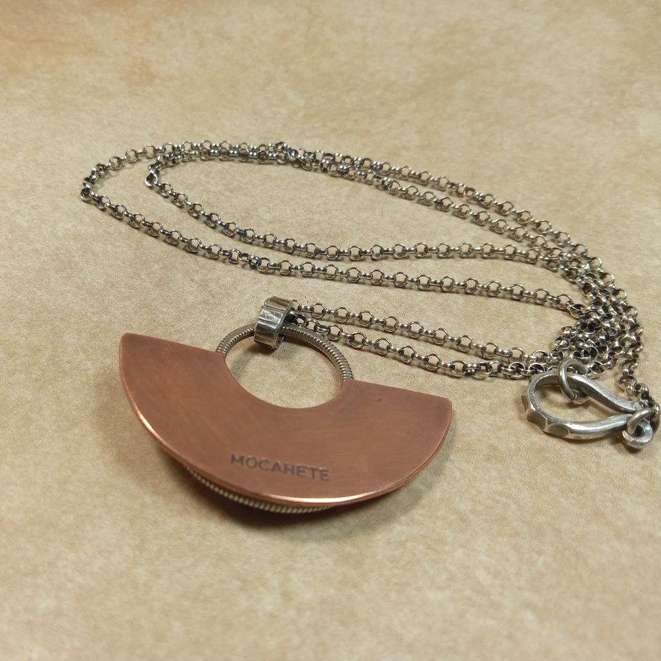 Reverse side of Handcrafted mixed metal lotus pendant in argentium silver and copper with lapis lazuli cabochon, showing Mocahete makers mark. Displayed on neutral background.