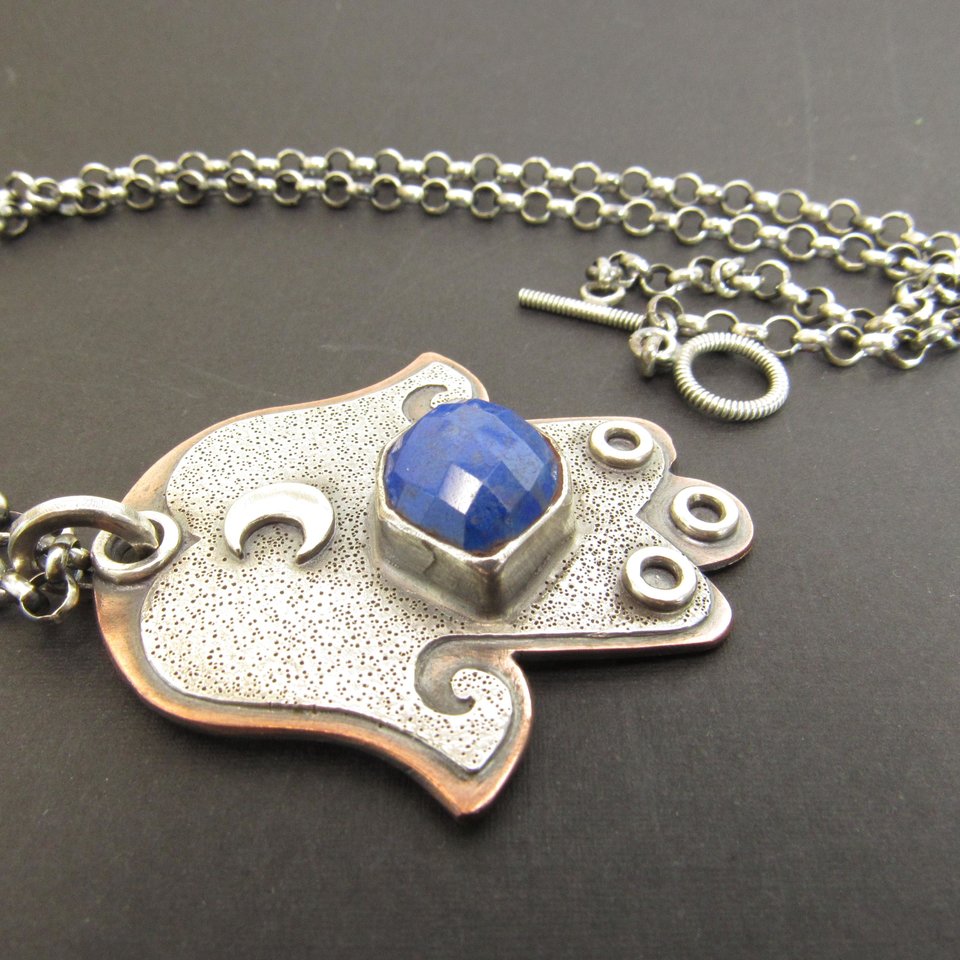 Mixed metal, copper and argentium sterling silver hamsa pendant necklace with lapis lazuli cabochon and crescent moon detail. 19 inch ss chain and findings. Hamsa is 1.5" wide x 1.75" tall. Displayed against dark background.
