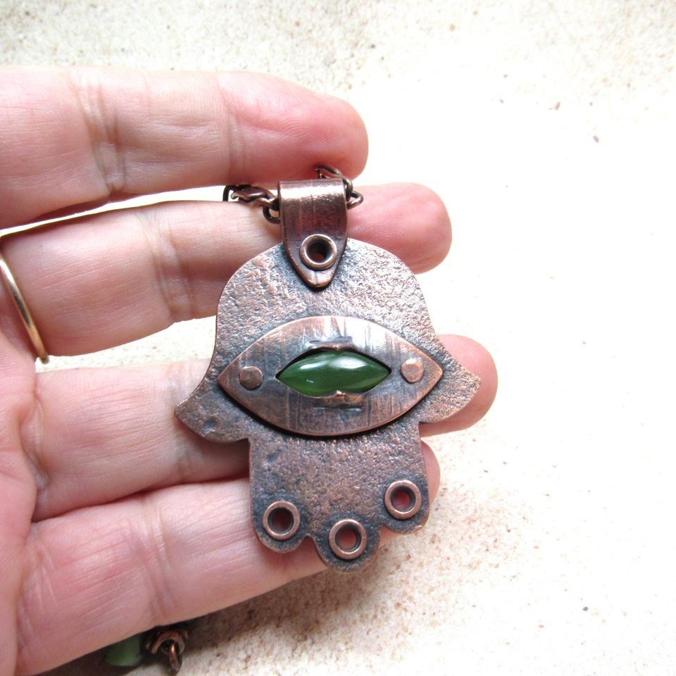Handcrafted copper, jade and chrysoprase hamsa pendant necklace. 2" tall x 1.5" wide, 24.5" copper scroll chain with 3 faceted chrysoprase beads on each side. All riveted design. Displayed against fingers of hand on neutral background.