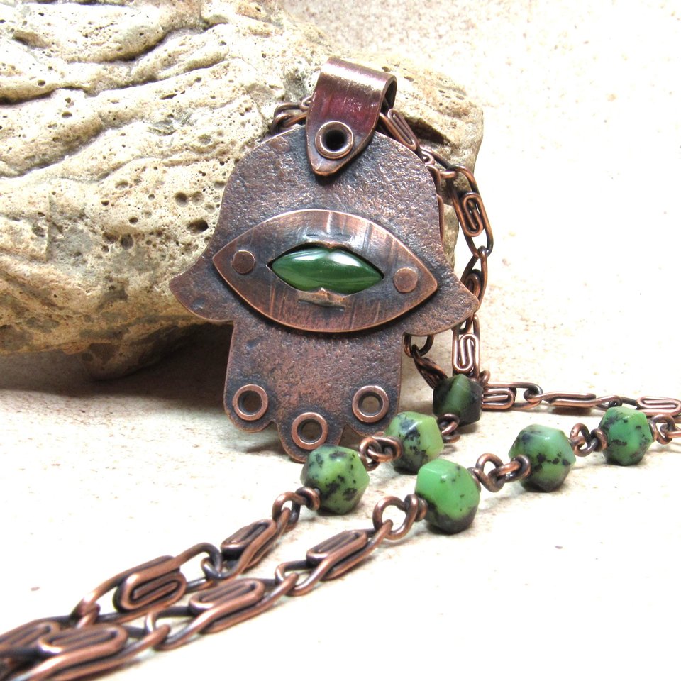 Up close photo of handcrafted copper, jade and chrysoprase hamsa pendant necklace. 2" tall x 1.5" wide, 24.5" copper scroll chain with 3 faceted chrysoprase beads on each side. All riveted design. Displayed against rock on neutral background.