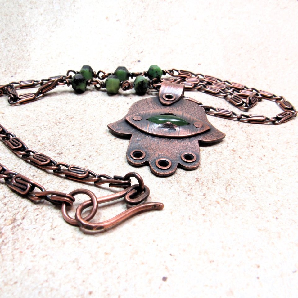 Handcrafted copper, jade and chrysoprase hamsa pendant necklace. 2" tall x 1.5" wide, 24.5" copper scroll chain with 3 faceted chrysoprase beads on each side. All riveted design. Up close of necklace hook closure. Displayed on neutral background.