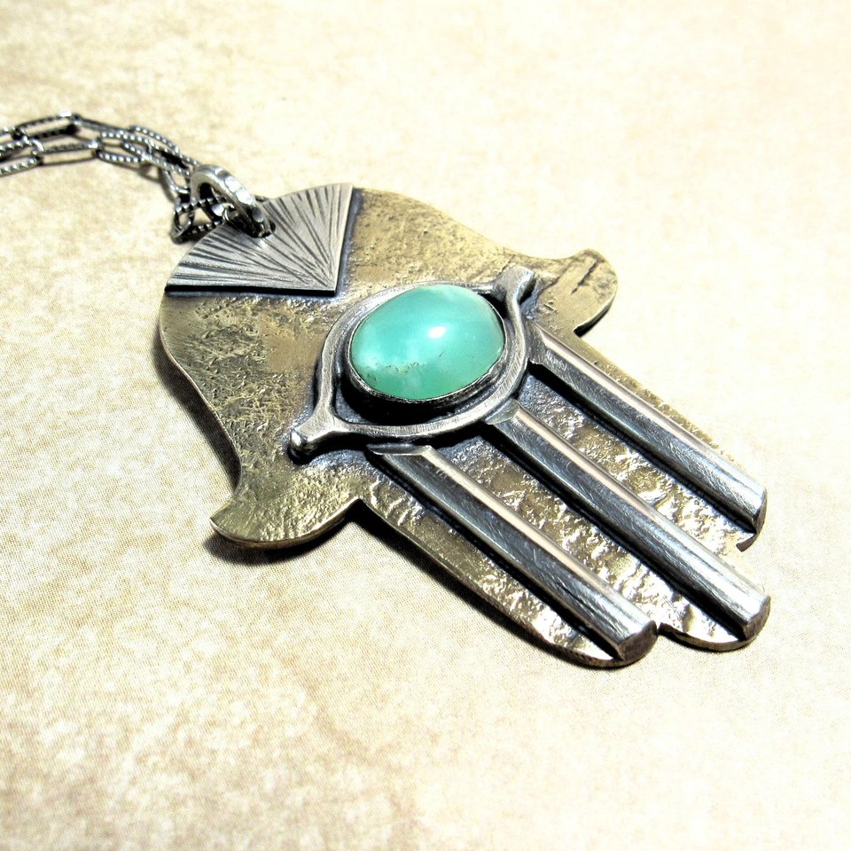 Bronze and silver hamsa pendant necklace with green stone displayed on neutral background. Pendant measures 1.5" wide x 2" long.