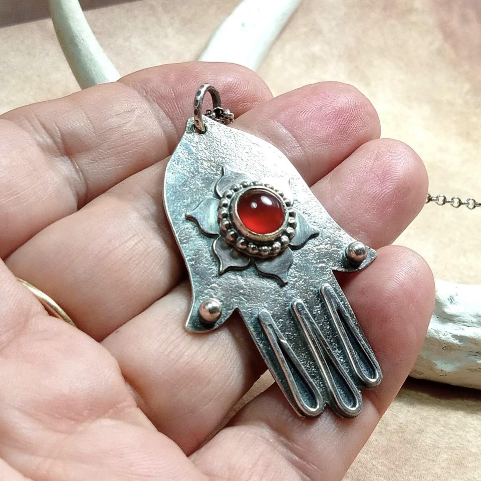 SS Hamsa pendant with carnelian, displayed in hand against antler and brown background.