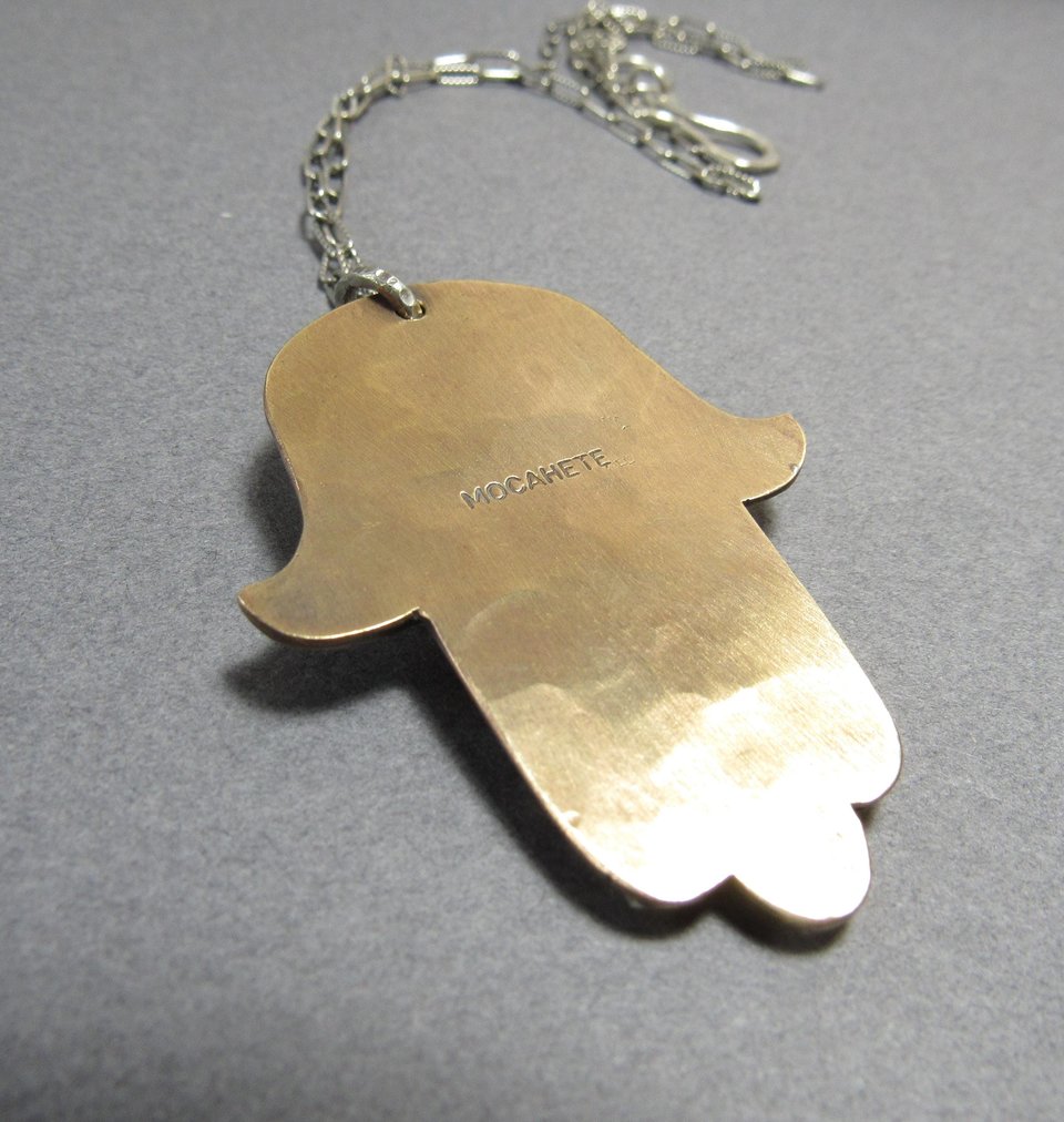 Reverse side of bronze and silver hamsa showing makers mark " Mocahete". Displayed on grey background.