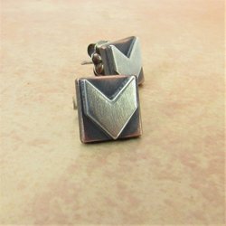 A pair of silver and copper earrings with a geometric arrow design. The earrings are square shaped with a brushed silver arrow in the center.