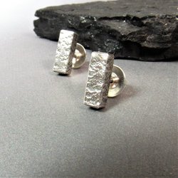 A pair of silver bar stud earrings with a textured finish. The earrings are on a gray surface with dark rock.