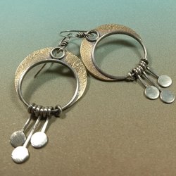 Artisan dangle earrings with swinging silver paddles beneath bronze crescent moons. Earrings by Mocahete photo on blue and brown background.