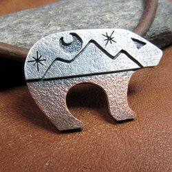 Bear pendant with the top half sculpted in silver with moon and star cutouts and the lower body in richly textured copper, joined harmoniously through traditional metalsmith techniques. The pendant is suspended on a sturdy 18-22 inch leather cord finished with our handmade Argentium silver findings for durability and comfort.Bear measures 1.25" long and is .75" tall. Displayed on brown leather against rock.